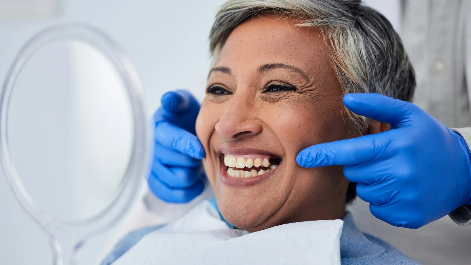Limpieza Dental en Tamarac: Qué Incluye la Primera Visita y Cada Cuánto Volver 1 Una paciente mayor sonríe y observa su sonrisa en un espejo de mano durante una cita dental