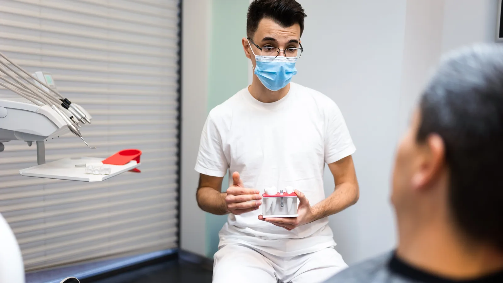 A dentist holds a small dental model while explaining treatment options to a seated patient in the chair