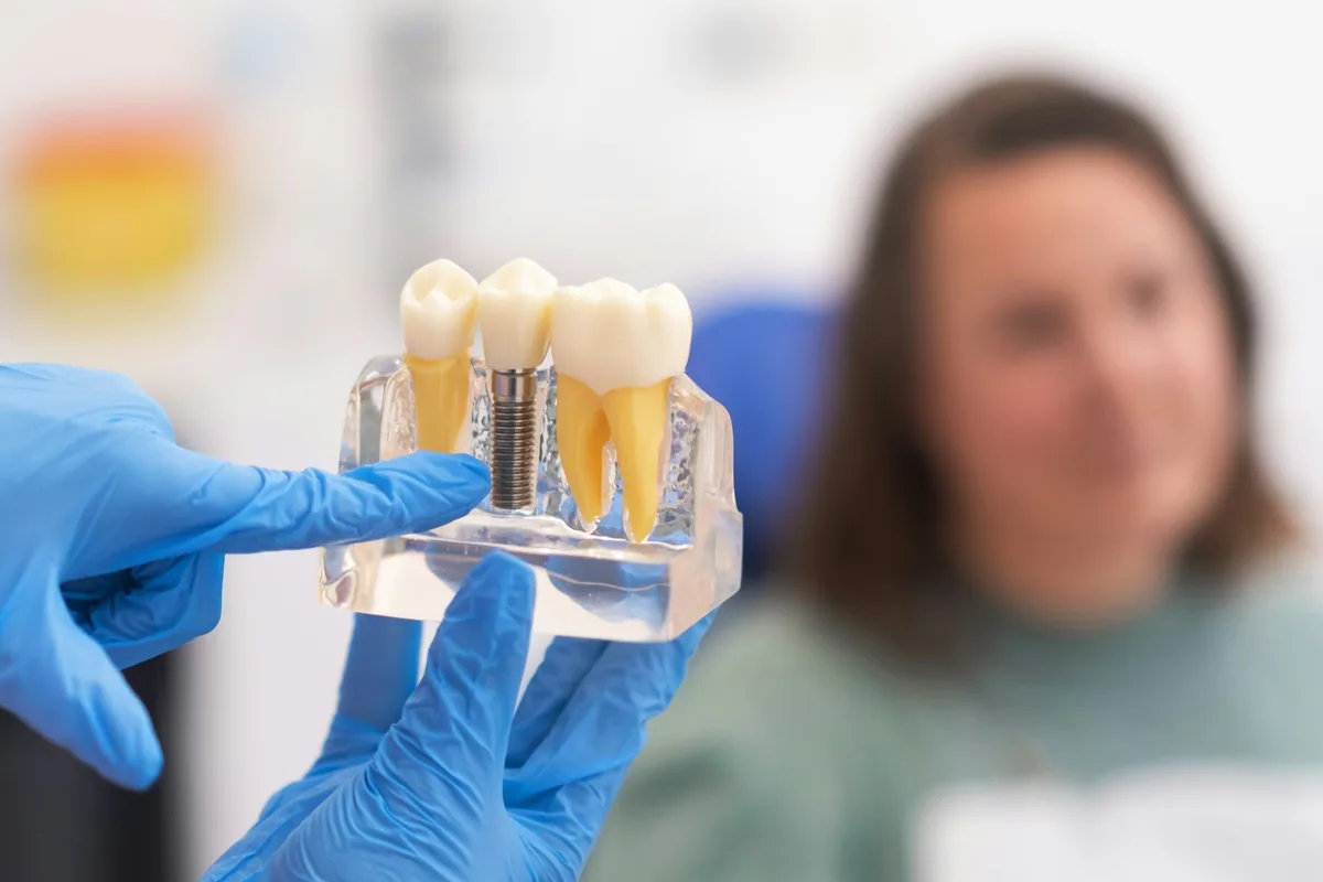 A dentist in blue gloves holds a clear model of a dental implant while talking with a seated patient