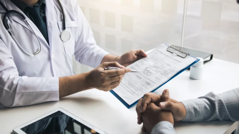A clinician points to a form on a clipboard across a desk while a patient listens with hands folded