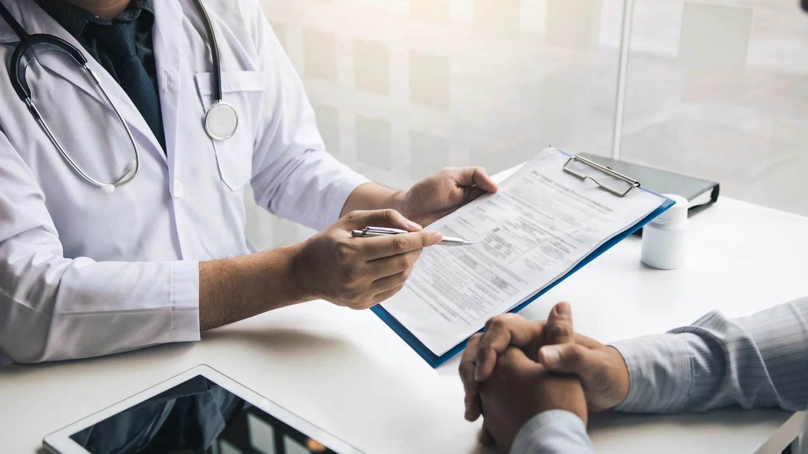A clinician points to a form on a clipboard across a desk while a patient listens with hands folded