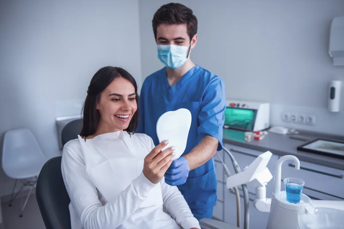 A patient in a dental chair smiles while holding a hand mirror as a clinician stands beside her
