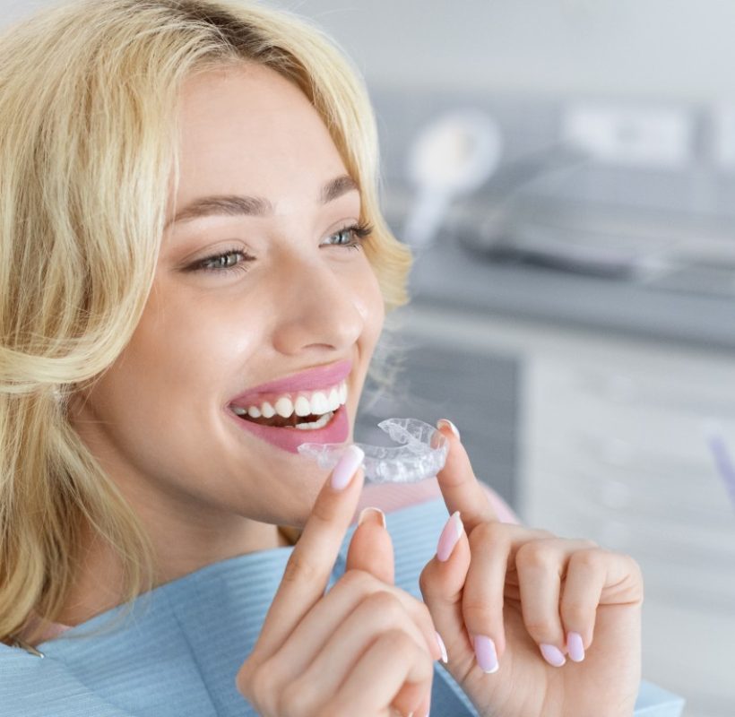 Invisalign orthodontics, modern dentistry concept. Happy young attractive blonde woman holding invisible braces or trainer while sitting at dental chair, closeup portrait, copy space